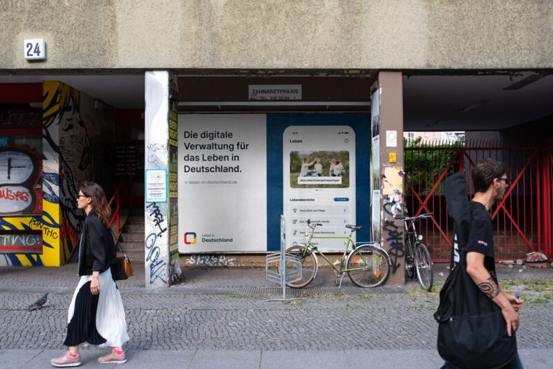 Straße mit Blick auf Gebäudewand, darauf ein Plakat in Zweiteilung, links Schriftzug
