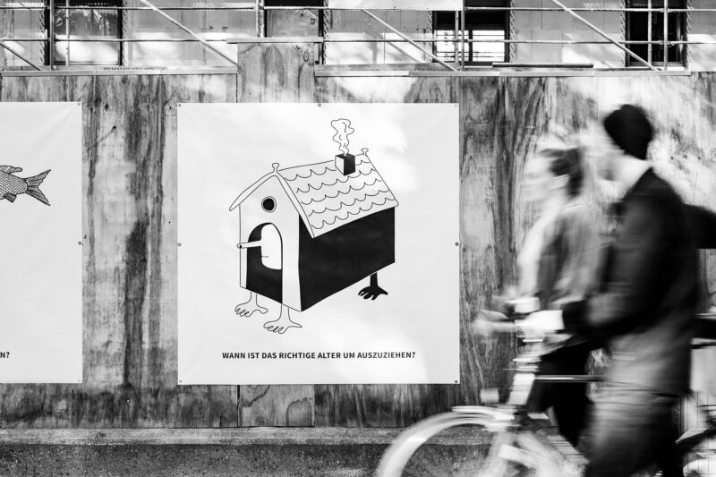 Bauzaun mit gedrucktem quadratischen Plakat, darauf eine Illustration mit einem Haus mit Beinen und dem Untertext